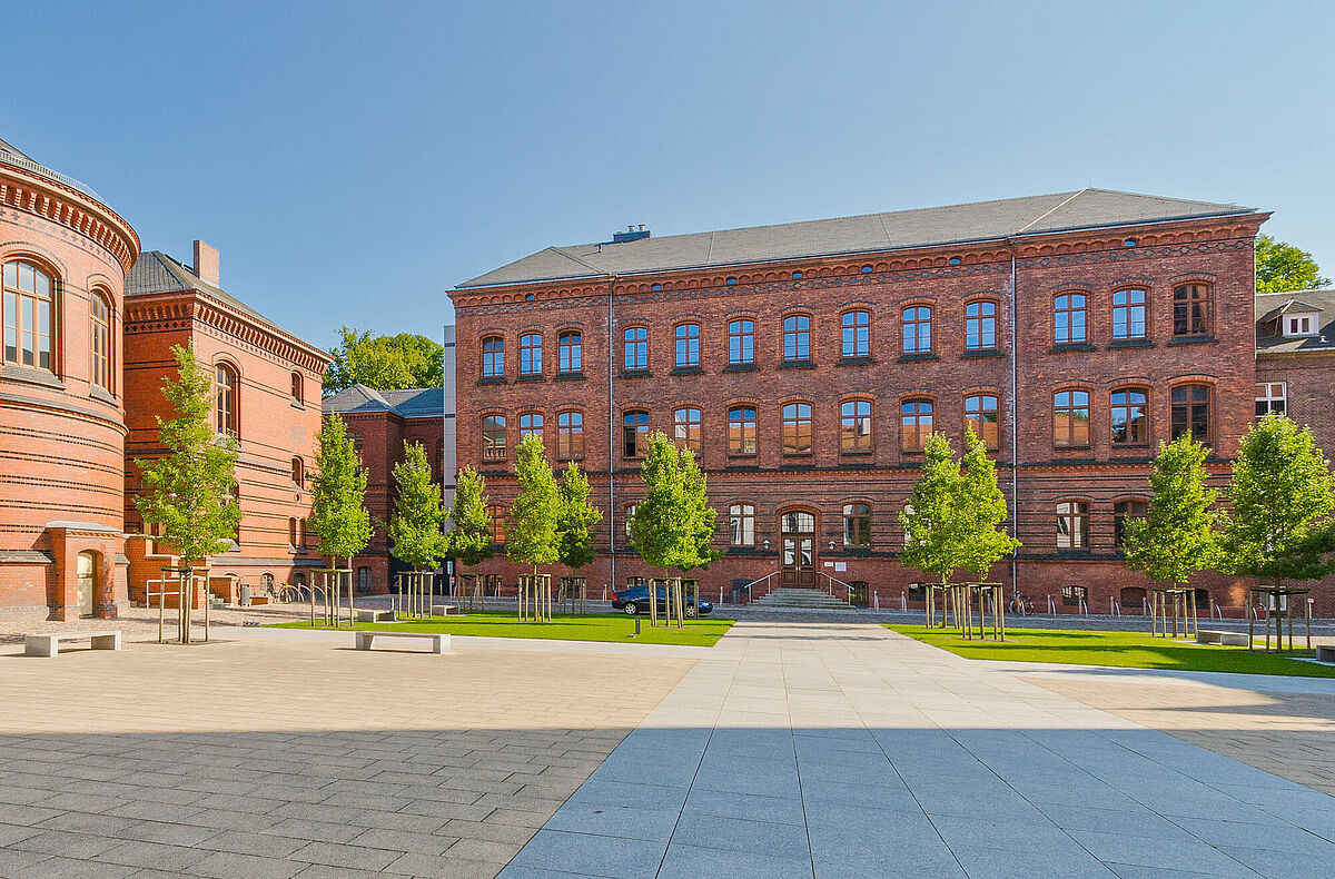 Außenansicht der backsteinernen ehemaligen Augenklinik hinter dem begrünten und sonnenbeschienen Innenhof des Historischen Campus vor blauem Himmel.
